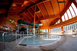 An indoor swimming pool with a slide, surrounded by large windows and a high ceiling.