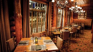 A cozy log cabin-style restaurant with wooden tables and chairs, large windows, and a chandelier.