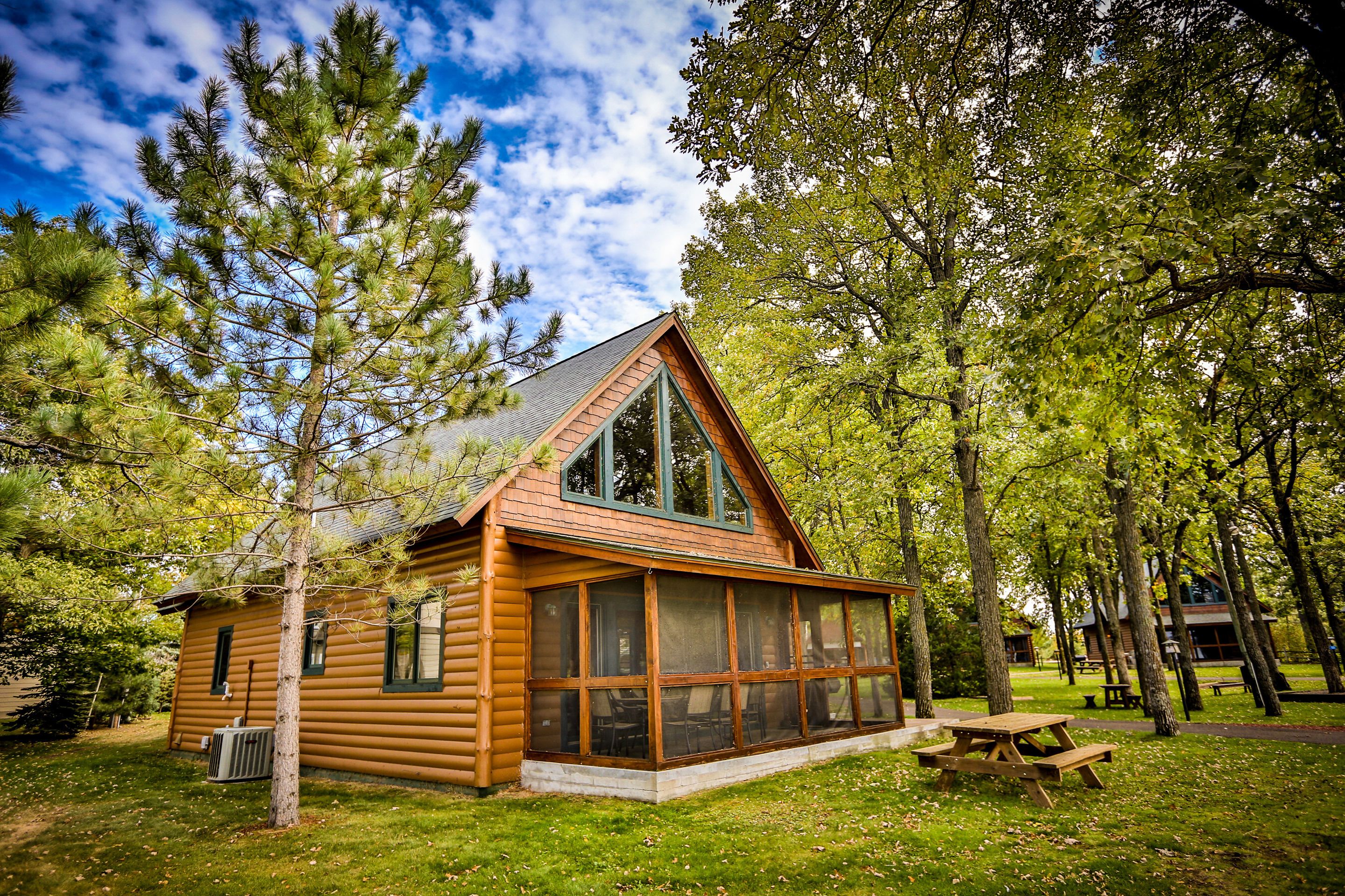 A log cabin with large windows and a picnic table in a wooded area.
