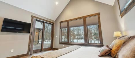 Un dormitorio con cama, ventilador de techo, televisor montado en la pared y ventanas amplias con vista a árboles.