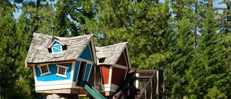 A treehouse play structure with a slide, surrounded by a grassy field and tall trees.