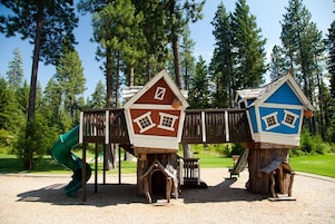 Un parque infantil con dos casitas de madera, un tobogán verde y un puente de madera.