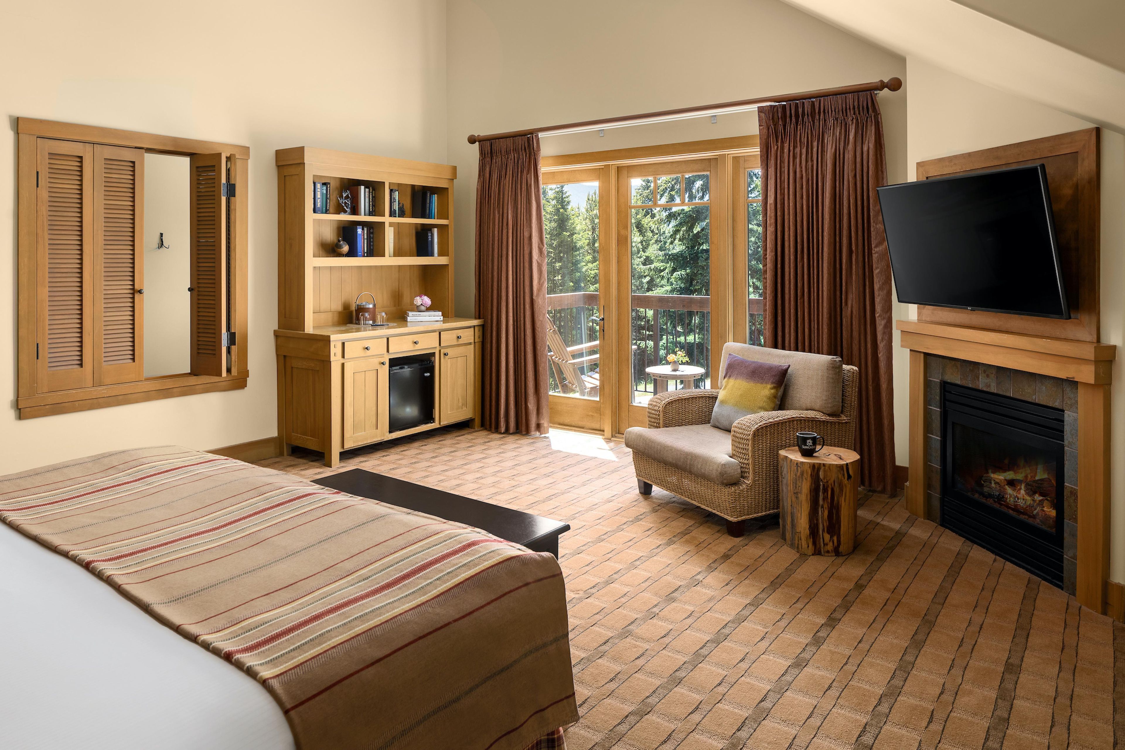 Une chambre d’hôtel comprenant un lit, une télévision, une cheminée, un balcon avec des rideaux et une armoire en bois remplie de livres.