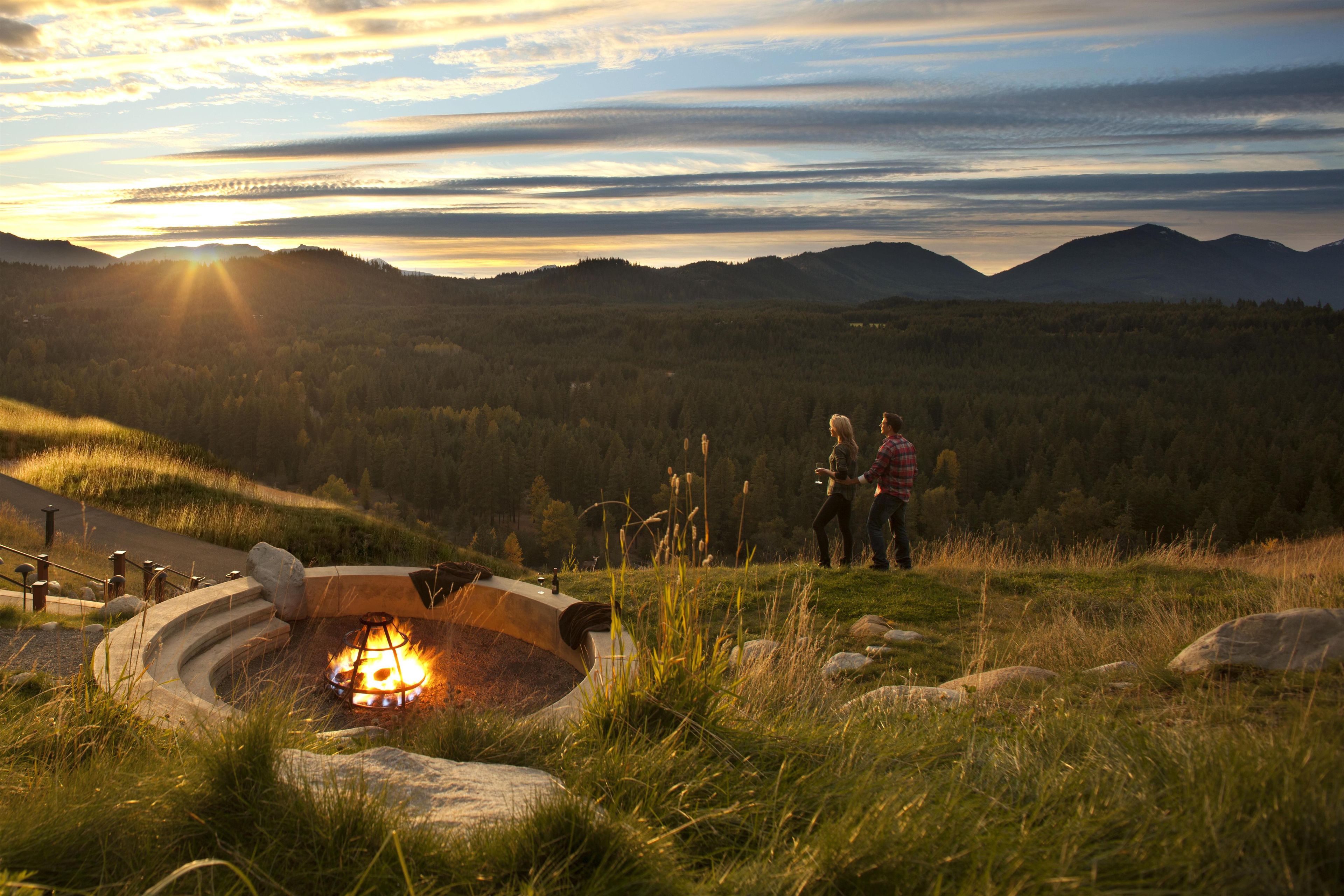 A campsite with a fire pit surrounded by rocks and grass, overlooking a forested valley with mountains in the distance.