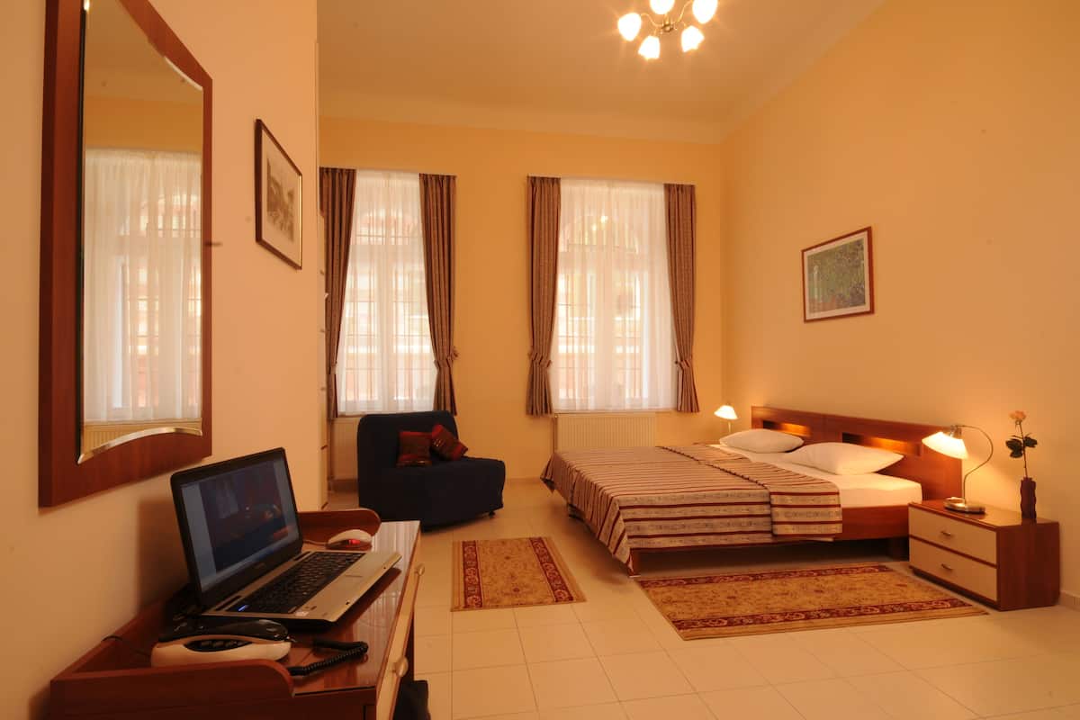 A hotel room with a bed, a desk with a laptop, a chair, a bedside table with a lamp, and a window with curtains.