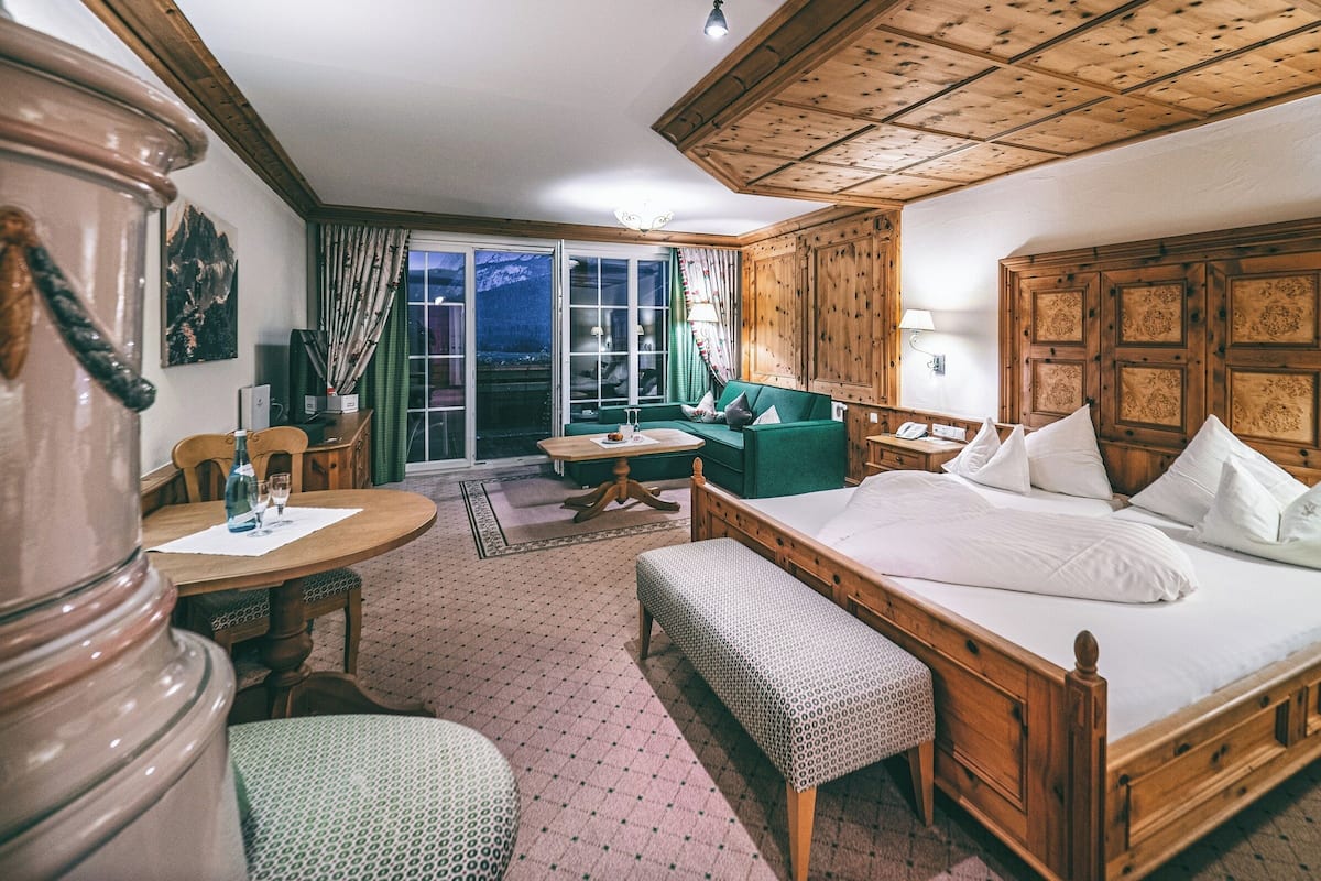 A hotel room with a wooden bed, a green sofa, a dining table with chairs, a small table with a bottle and glasses, and a large window with a view of mountains.