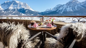 A table set outdoors with a mountain view, wicker chairs, and a bottle of wine.