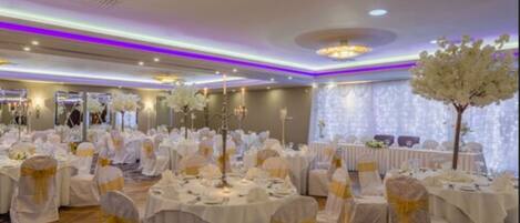 Un salón de banquetes con mesas redondas, manteles blancos y sillas con cintas doradas.