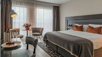 Una habitación de hotel con cama, escritorio con lámpara, silla, pequeña mesa con florero y una botella de champán.