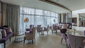 Un vestíbulo amplio con techo curvo, ventanas grandes con cortinas translúcidas y diversas opciones de asientos, incluyendo sofás y sillones.