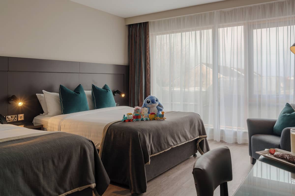Ein Hotelzimmer mit zwei Betten, einem Sofa, einem Tisch mit einer Kuchengarnitur und einem Glas Milch, einer Lampe und einem Fenster mit durchsichtigen Vorhängen.