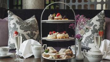 Un expositor de varios niveles con pasteles y sándwiches, acompañado de tazas de té y un florero con flores.