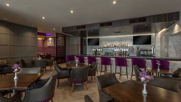 A modern bar area with a long counter, multiple seating options, and a television screen.