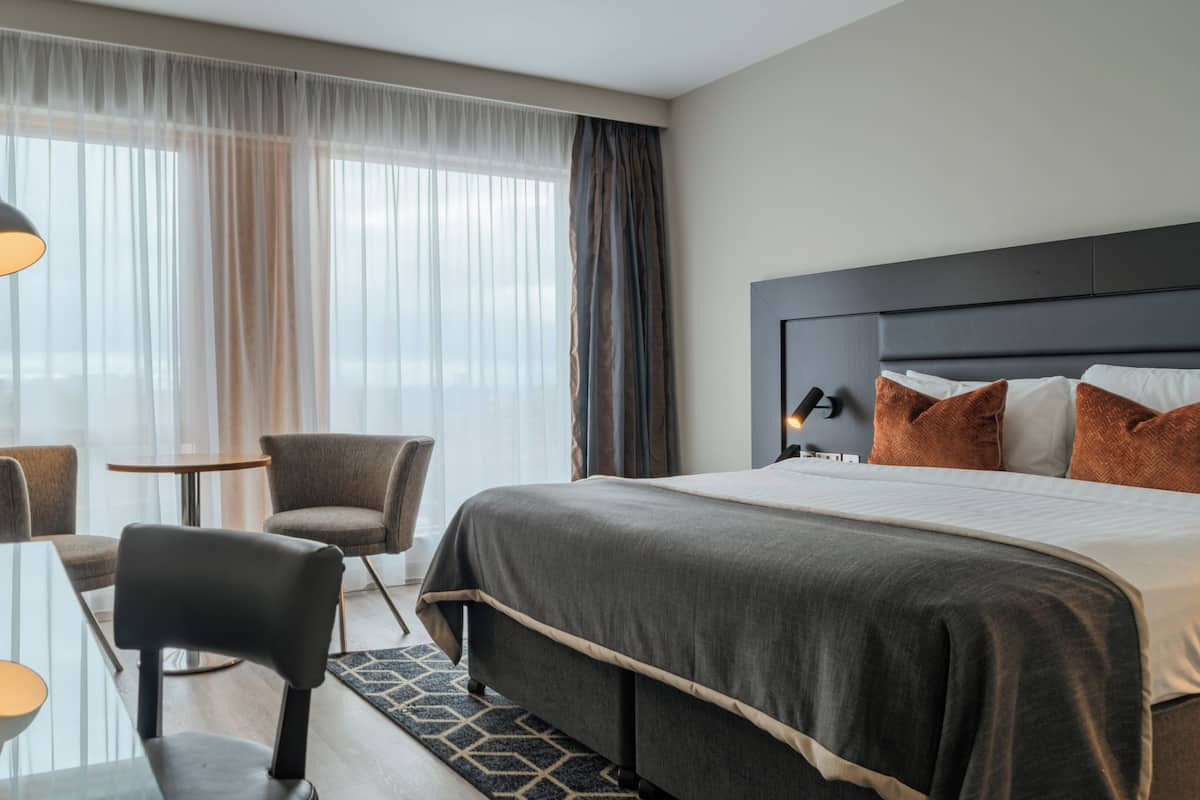 Ein Hotelzimmer mit Bett, zwei Stühlen, einem Tisch, einer Lampe und Blick durch transparente Vorhänge.
