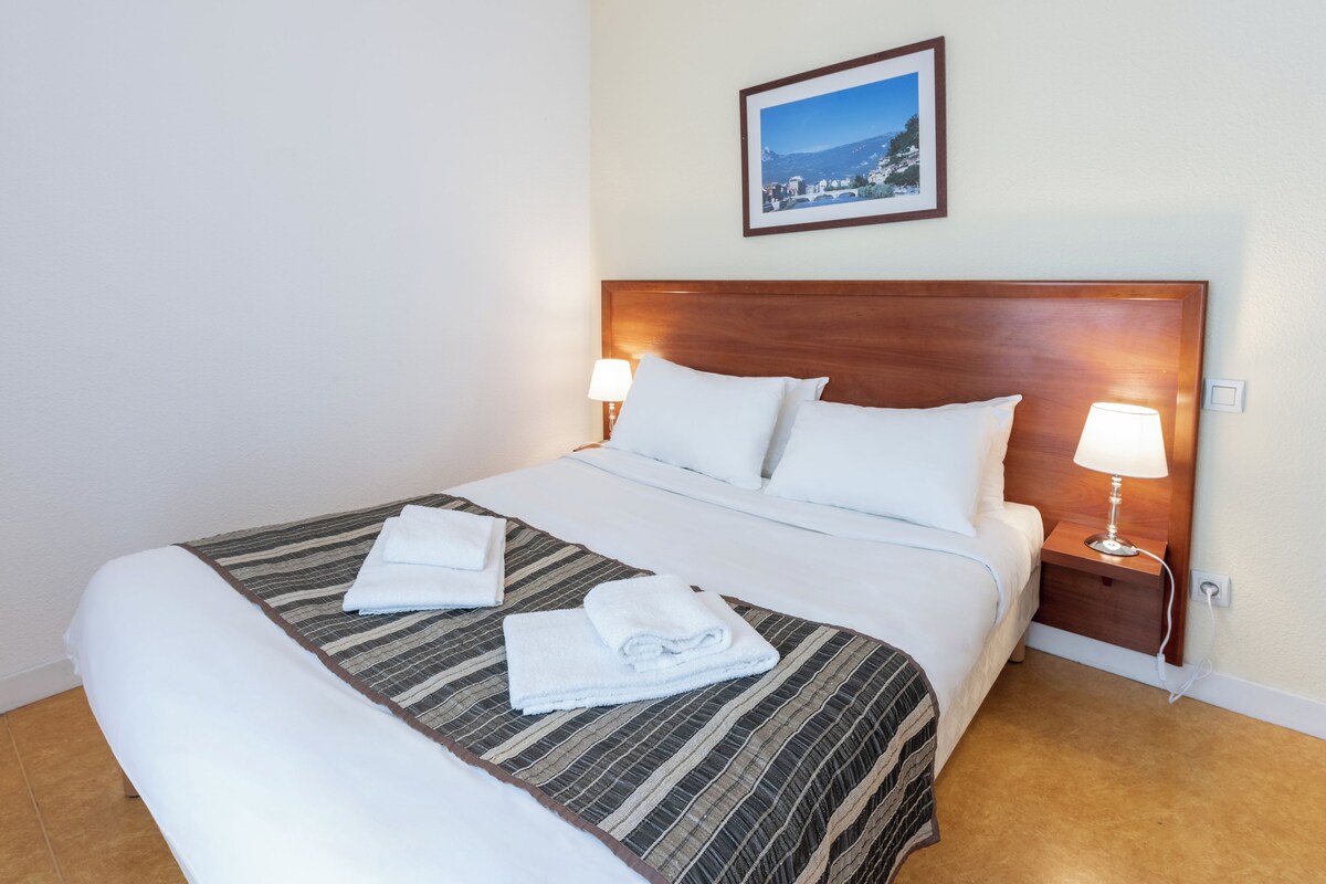 A neatly made bed with a striped bedspread, two pillows, and two folded towels. There is a framed picture on the wall above the bed.
