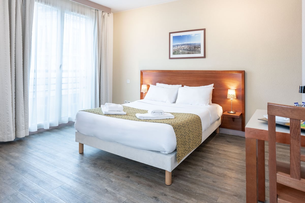 A hotel room with a large bed, two bedside lamps, a wooden headboard, a framed picture on the wall, and a wooden table with a chair.