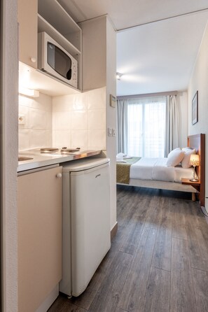 A compact hotel room with a kitchenette, a bed with white linens, a bedside lamp, and a view of the city through the window. - Residhotel Grenette (Grenoble)