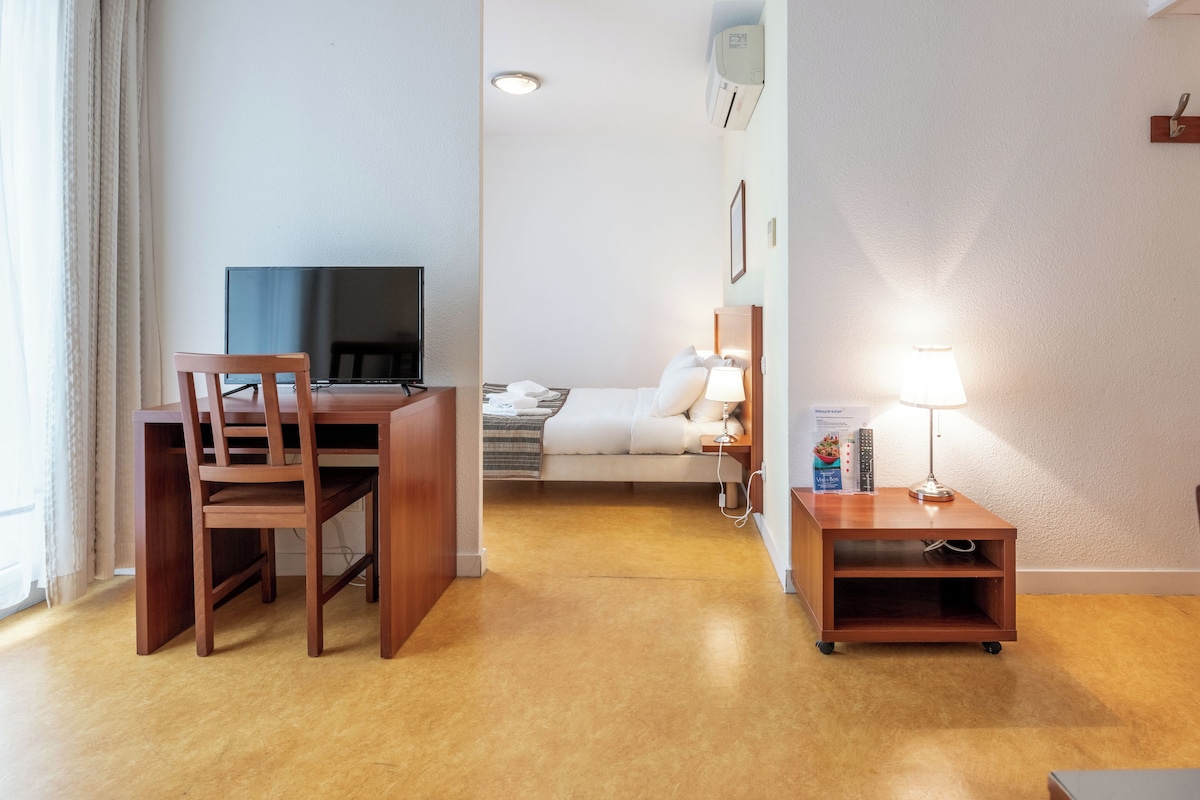 Hotellihuone, jossa on puinen työpöytä, sänky, televisio ja yöpöytälamppu.