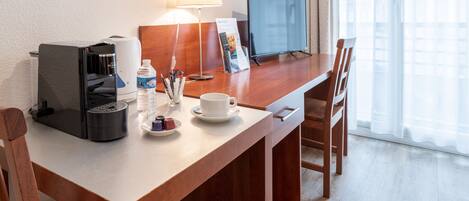 A hotel room with a wooden desk, a coffee maker, a water bottle, a cup, a lamp, and a TV.