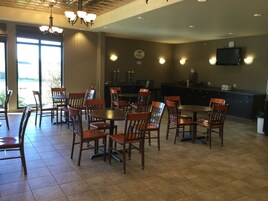 A spacious dining area with multiple wooden tables and chairs, a kitchen area with a flat-screen TV, and large windows allowing natural light.