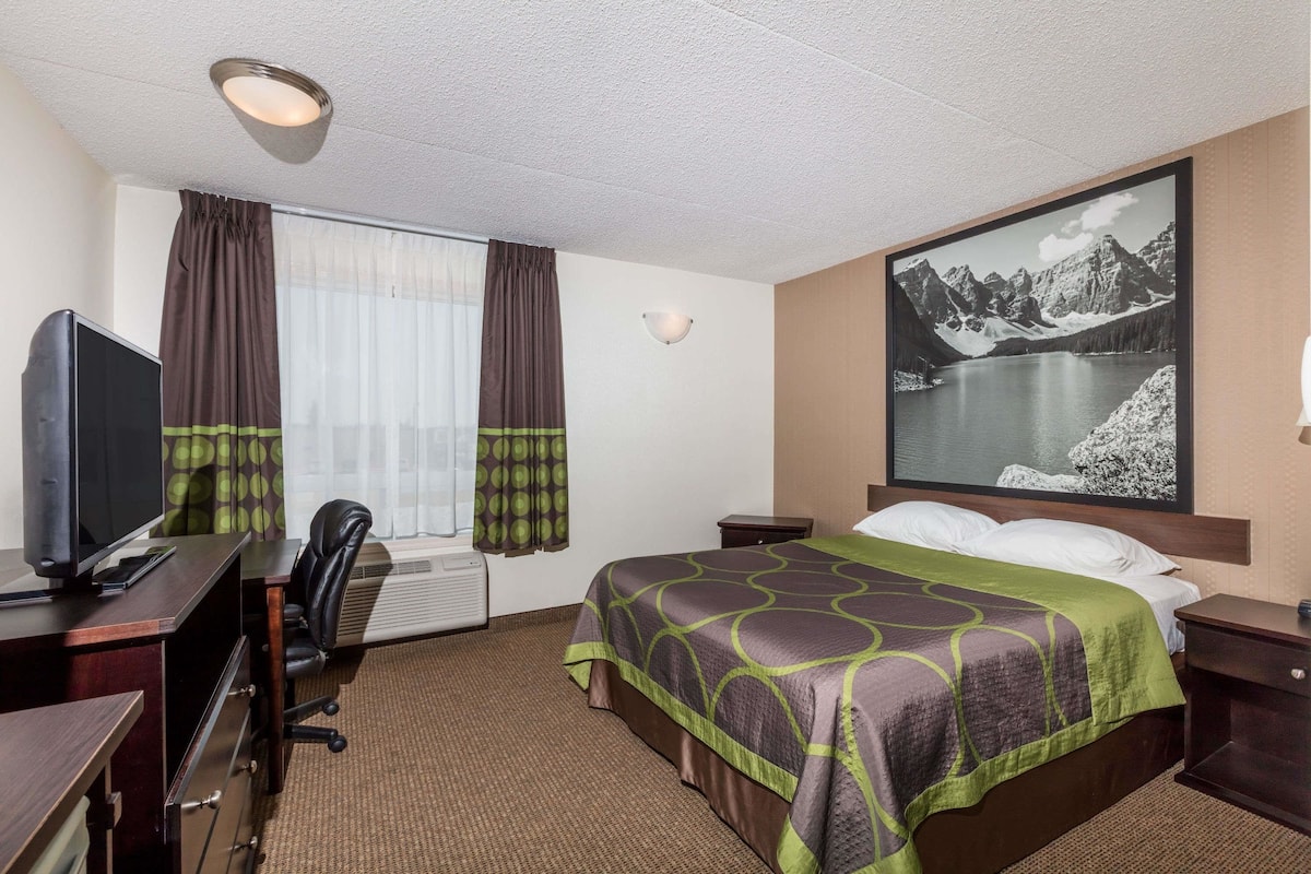 Ein Hotelzimmer mit Bett, Schreibtisch inklusive Computer, Fernseher und großem Fenster mit Vorhängen.