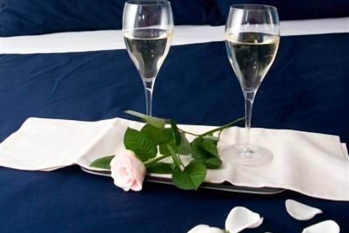 A bed with two glasses of white wine, a rose, and white flower petals on a navy blue bedspread.