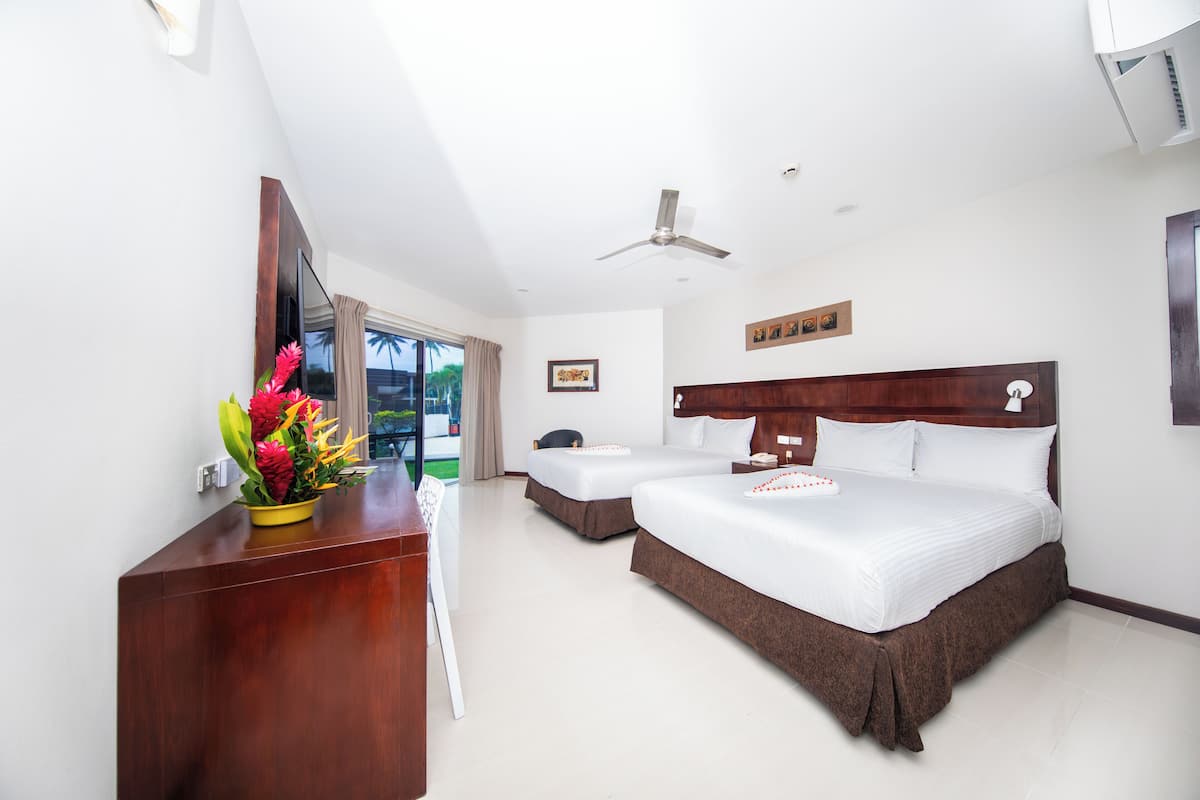 A hotel room with a large bed, a smaller bed, a desk with a vase of flowers, a ceiling fan, and a view of palm trees.