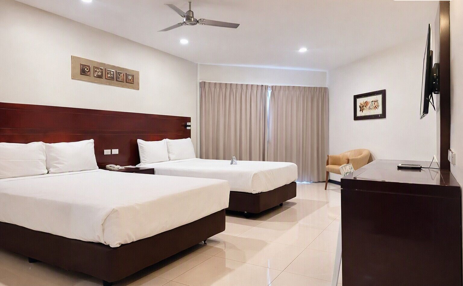 A hotel room with two beds, a desk, a chair, a television, and a ceiling fan.