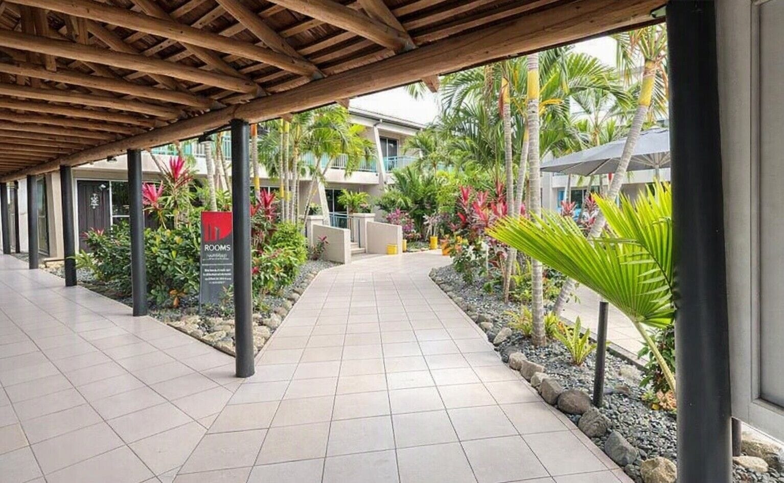 Een overdekte doorgang met betegelde vloer, omringd door tropische planten en palmbomen, die naar een gebouw leidt met een bord waarop 'ROOMS' staat.
