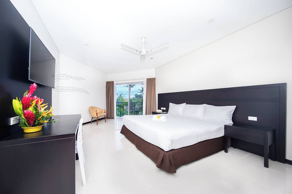 A modern hotel room with a large bed, a flat-screen TV, a desk, and a balcony with curtains.