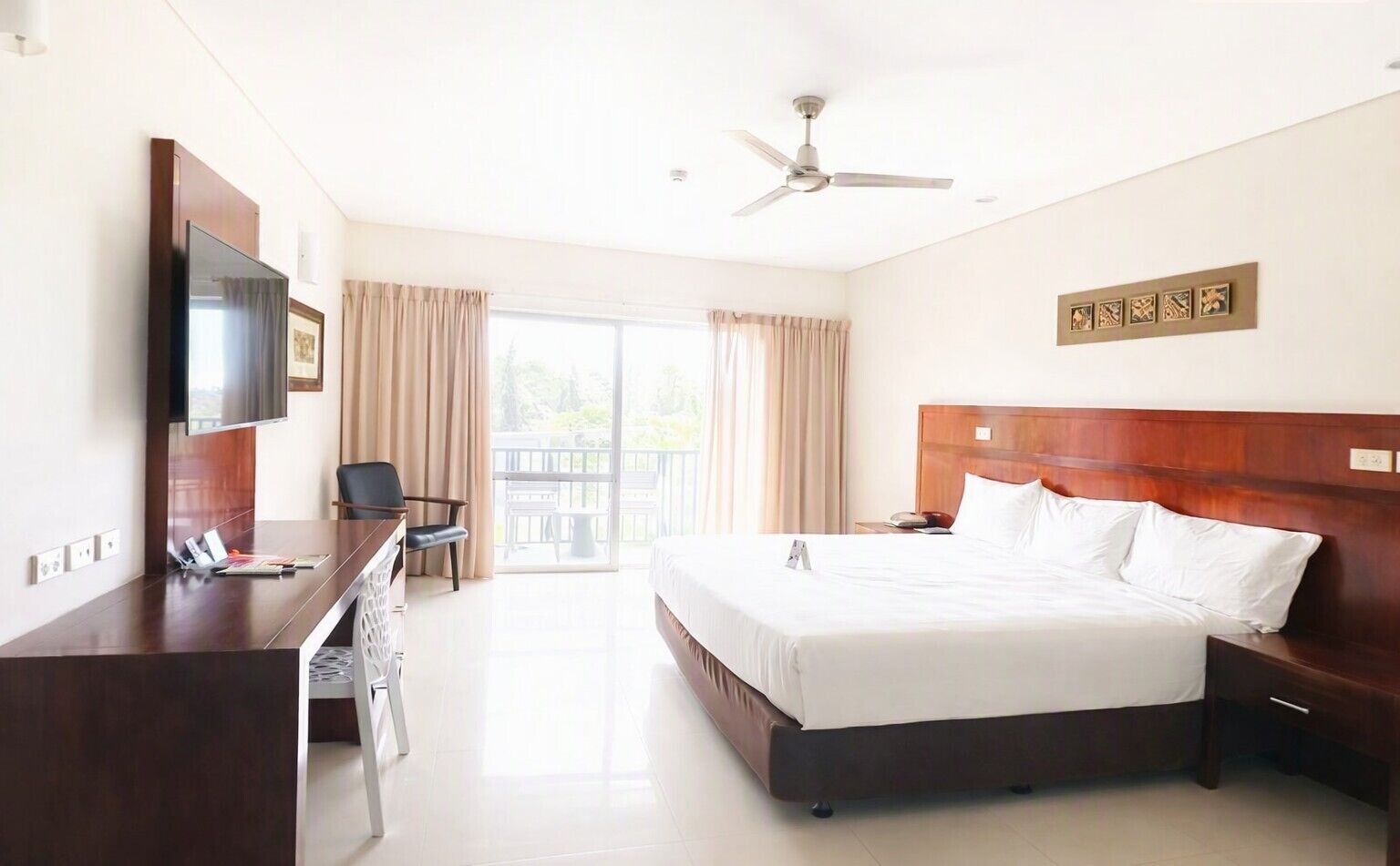 A hotel room with a bed, desk, chair, television, and a balcony with curtains.