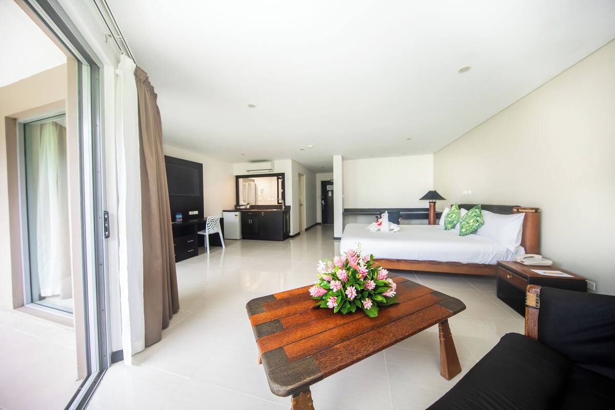 A modern hotel room with a bed, a wooden coffee table with flowers, a black sofa, a desk, and a TV.