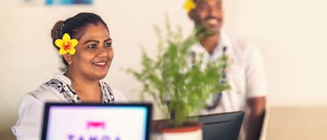 Una persona sentada en un escritorio con una computadora mostrando 'TAMOA' en la pantalla, una planta en maceta y otra persona al fondo.