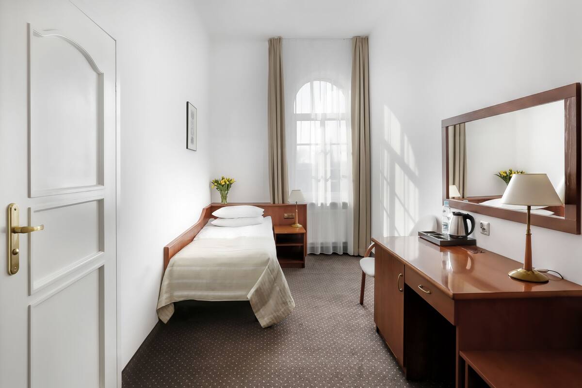 Camera d'albergo con un letto, una scrivania, una sedia, uno specchio, una finestra con tende e una piccola pianta.