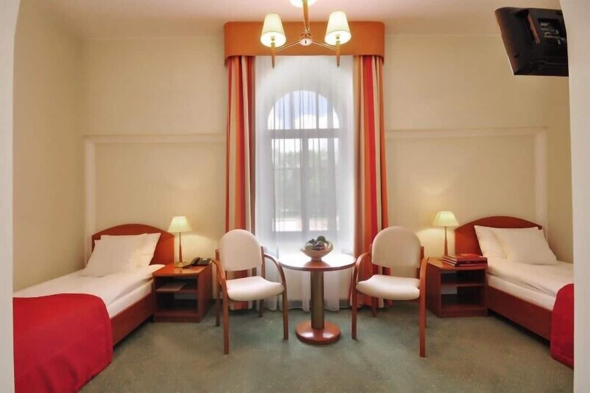 Una camera d'albergo con due letti singoli, un tavolino con sedie, una finestra con tende e un lampadario.