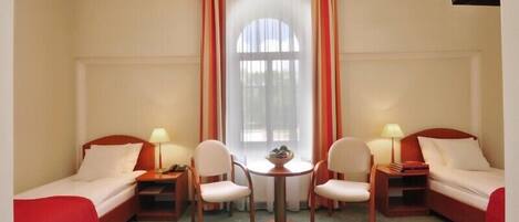A hotel room with two single beds, a small table with chairs, a window with curtains, and a ceiling light fixture.