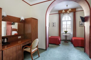 Une chambre d’hôtel avec un bureau en bois, une chaise, deux lits simples, une petite table avec un vase et une grande fenêtre en arc de cercle.