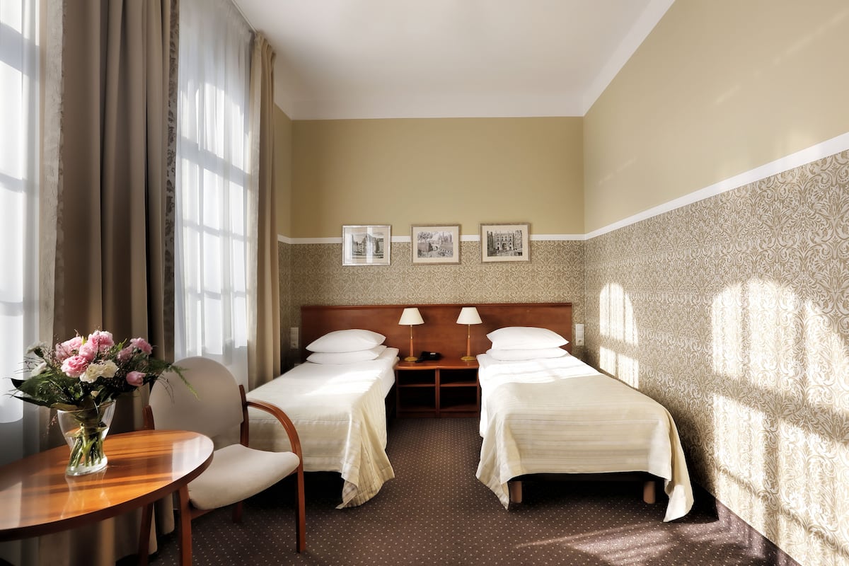 Camera d'albergo con due letti singoli, un tavolo in legno con un vaso di fiori, una sedia e un comodino con lampade.