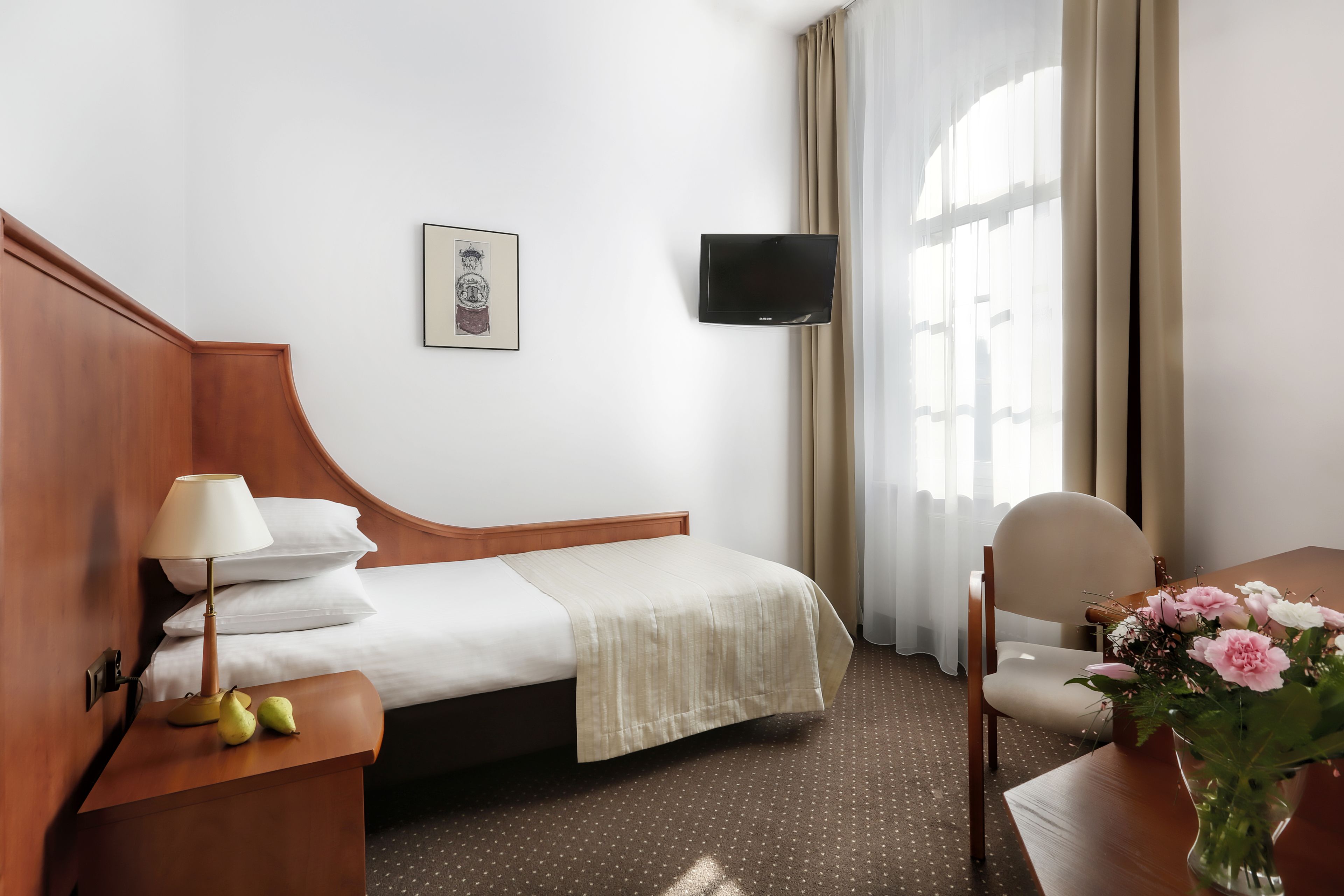 Une chambre d’hôtel comprenant un lit, une table de chevet avec une lampe, un téléviseur fixé au mur, une chaise et un vase avec des fleurs.