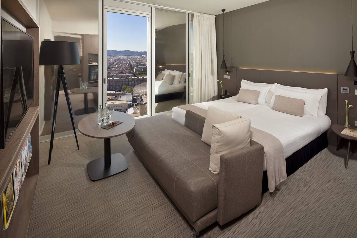Quarto de hotel moderno com uma cama grande, um sofá, uma mesa pequena e vista para a cidade.