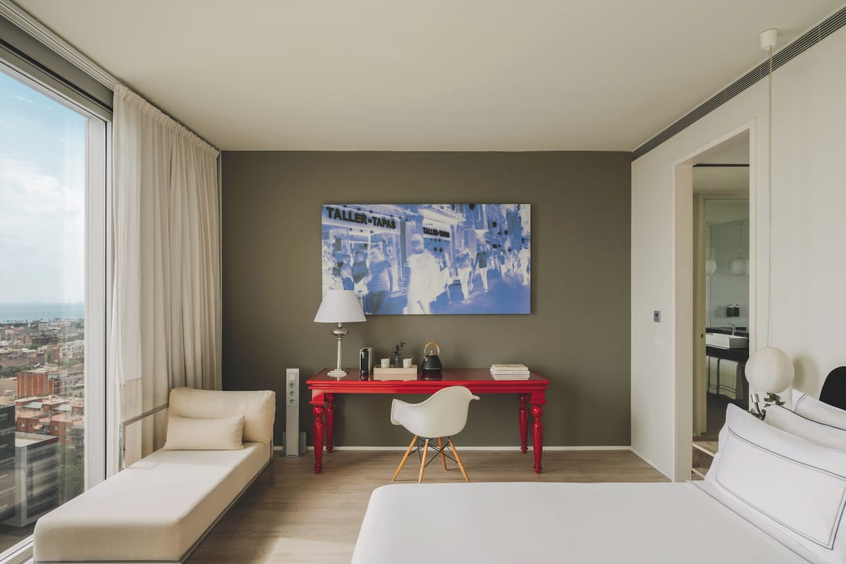 Quarto de hotel moderno com uma grande janela, uma secretária vermelha, uma cadeira branca, uma cama e vista para a cidade.