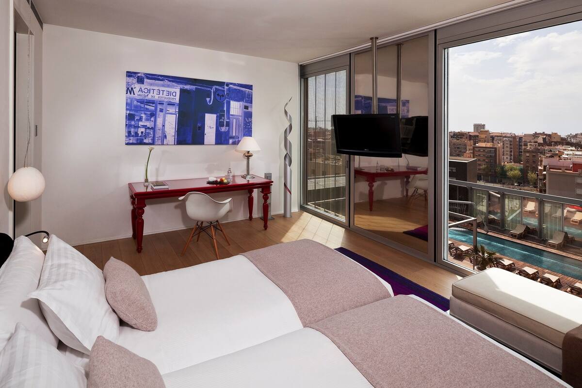 Quarto de hotel com cama, escrivaninha, cadeira, televisão e varanda com vista para a piscina.