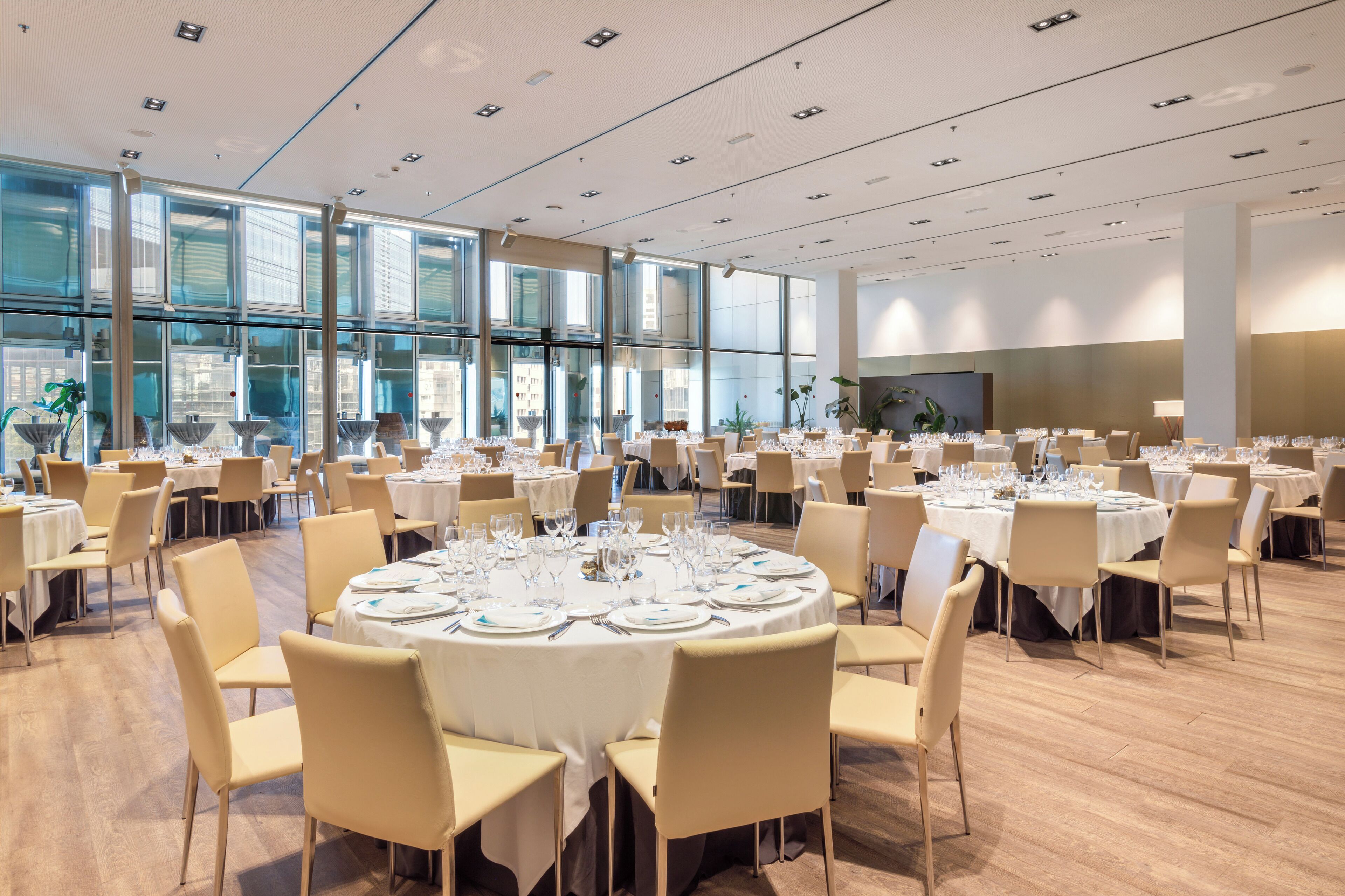 A spacious banquet hall with multiple round tables set for dining, large windows providing natural light, and a modern interior design.