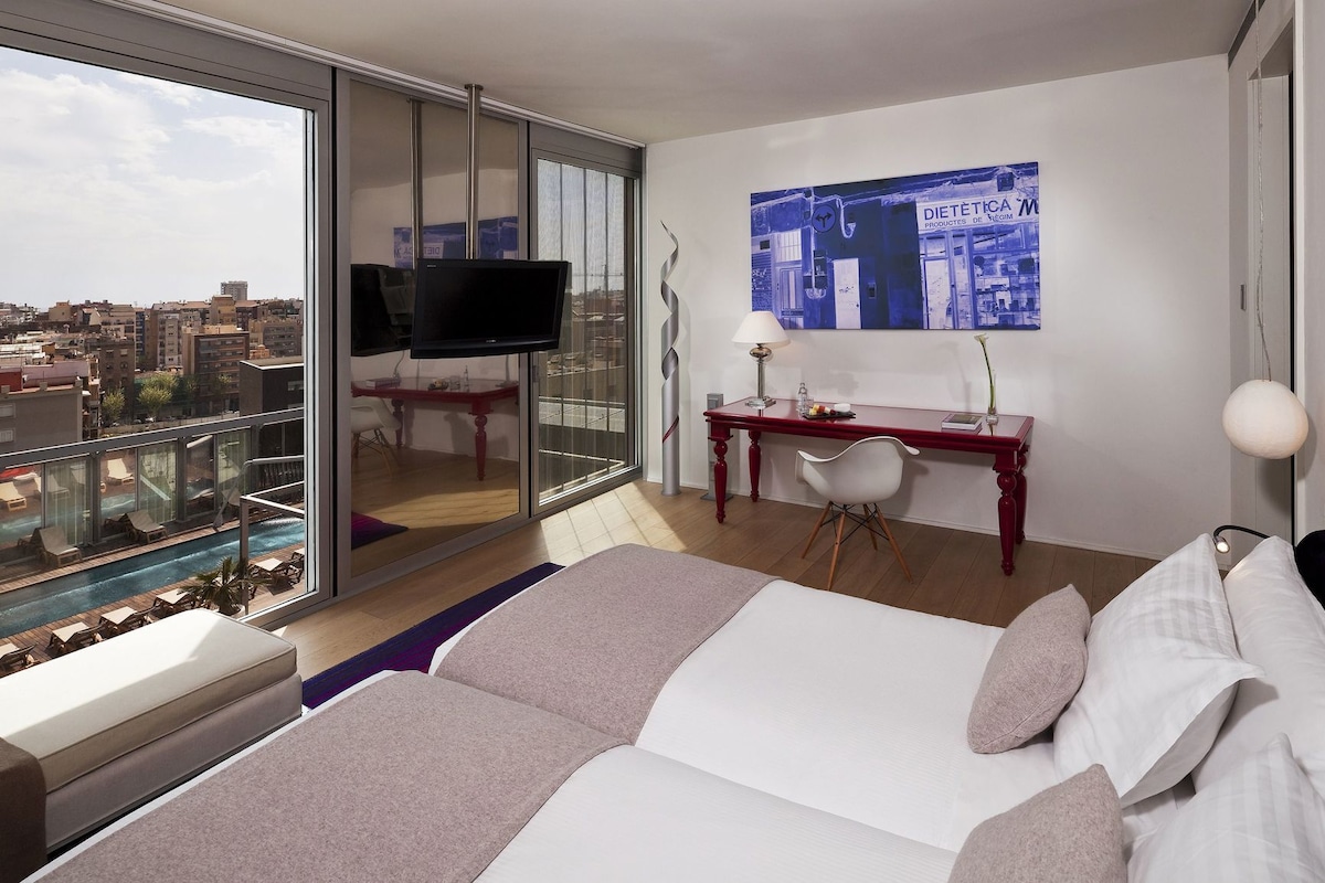 A hotel room with a bed, a desk, a TV, a city view, and a pool.