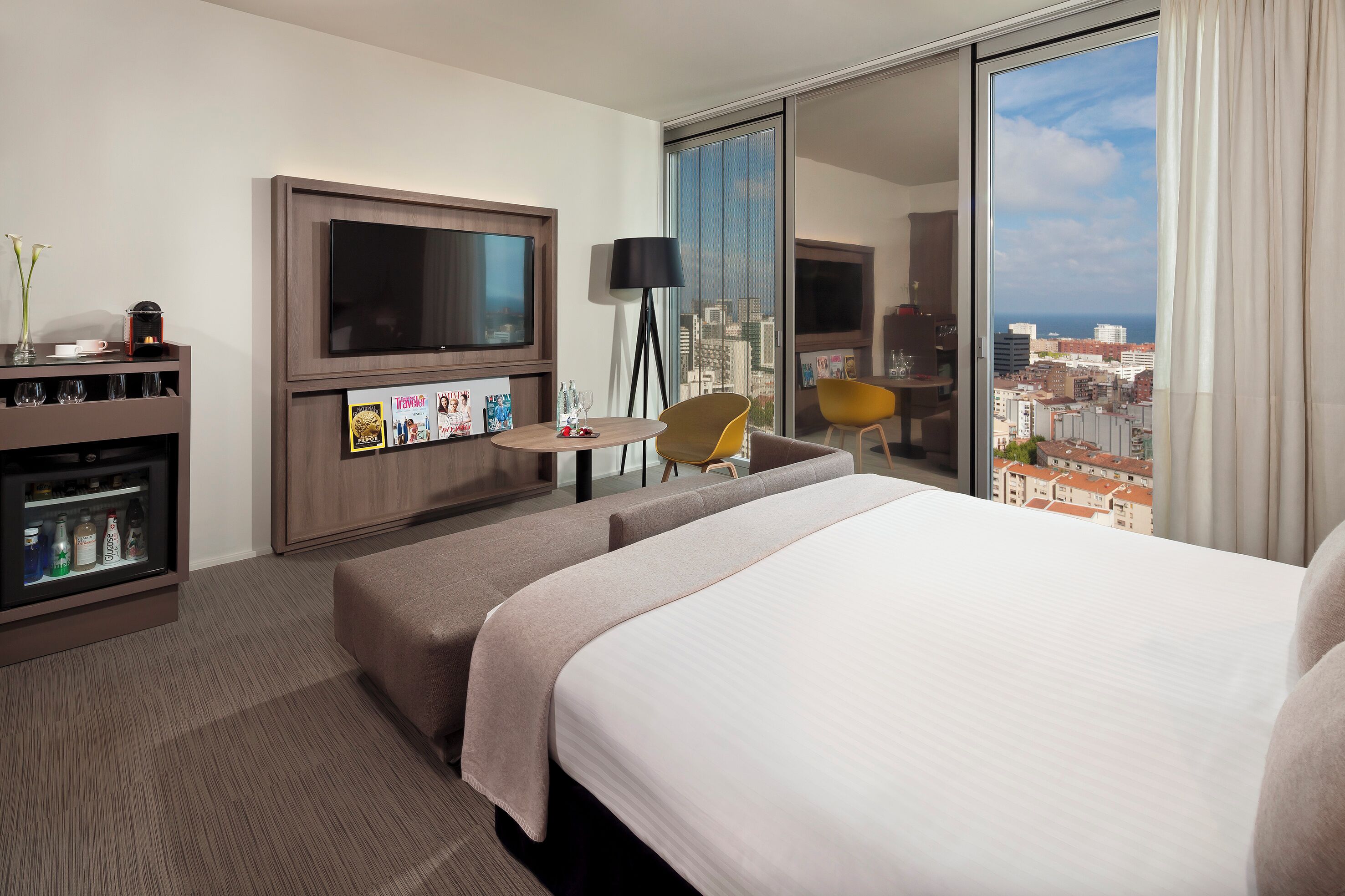Ein Hotelzimmer mit einem großen Bett, einem Fernseher, einem Schreibtisch mit Lampe und Blick auf Stadt und Meer.