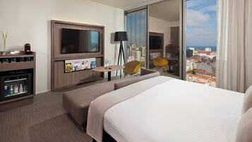 Ein Hotelzimmer mit einem großen Bett, einem Fernseher, einem Schreibtisch mit Lampe und Blick auf Stadt und Meer.