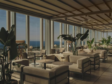 A spacious rooftop lounge with large windows, comfortable seating, and potted plants.. Melia Barcelona Sky