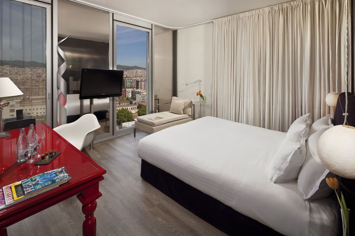 A hotel room with a bed, a TV, a desk with a magazine, a chair, and a view of the city.