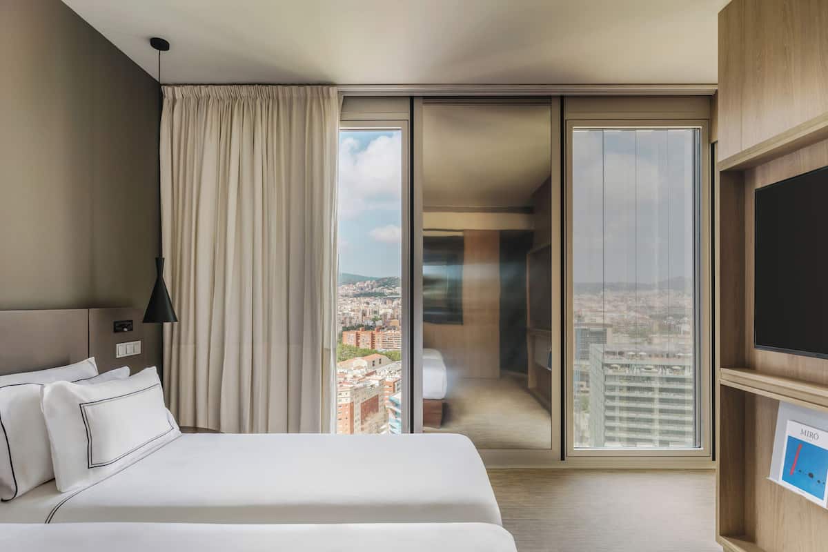 Quarto de hotel com cama, televisão, janela ampla com vista para a cidade e porta deslizante.
