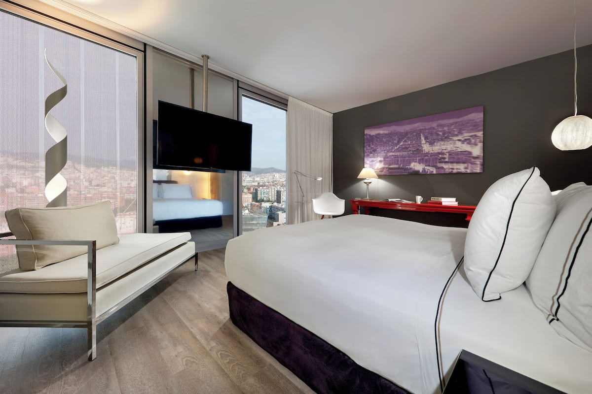 Quarto de hotel moderno com uma cama grande, um sofá, uma televisão e vista para a cidade.